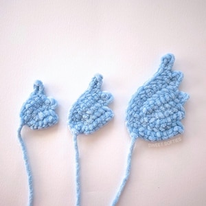 May include: Three light blue, crocheted wing shapes of varying sizes are displayed against a white background. The wings have a soft, textured appearance and are connected to long, thin strands of yarn. The text "SWEET SOFTIES" is visible on the largest wing.