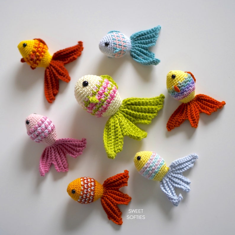 Puede incluir: Colecci&oacute;n de siete peces dorados de ganchillo en varios colores, incluyendo naranja, amarillo, rosa, azul y verde. Cada pez tiene un dise&ntilde;o detallado y texturizado y ojos de cuentas negras. La imagen est&aacute; sobre un fondo blanco.