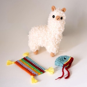 May include: A handmade, white crochet llama with a fluffy texture, standing near a colorful, rectangular rug and a small blue hat with red trim. The llama has brown ears and feet. The rug has yellow tassels.