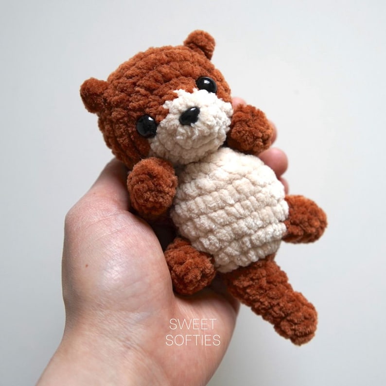 May include: A small crocheted stuffed animal in the shape of an otter. The otter is brown and white with black eyes. The otter is being held in a person's hand.