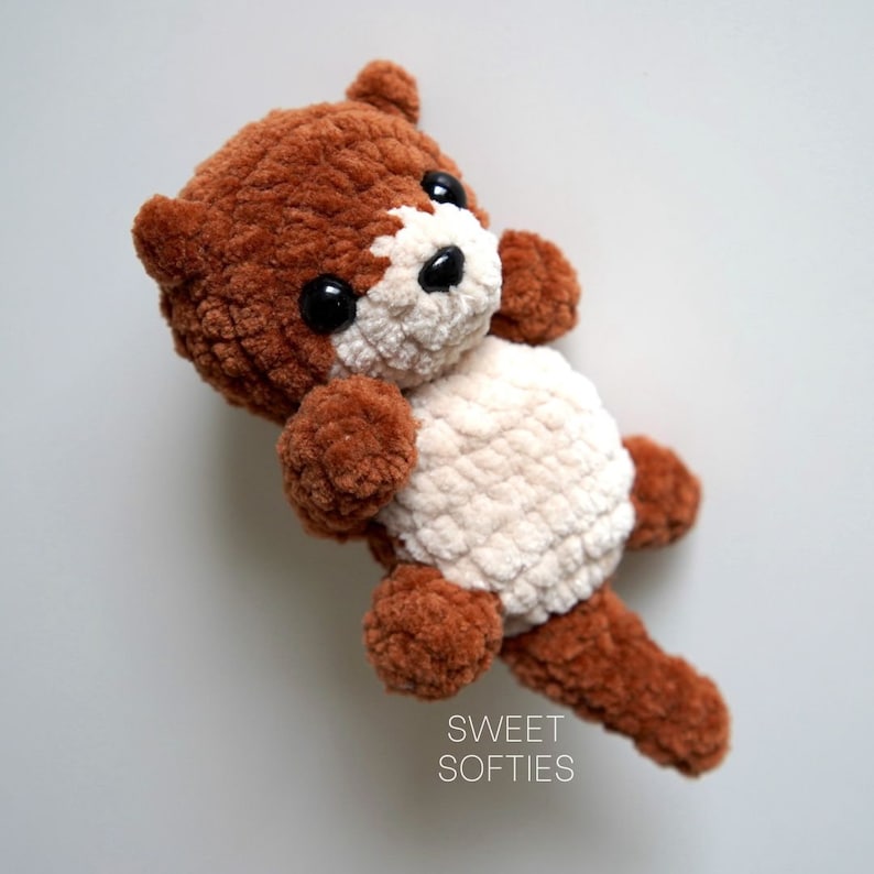 May include: A brown and cream crocheted stuffed animal with black eyes. The animal has a long tail and is lying on its back. The text "SWEET SOFTIES" is printed below the animal.