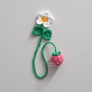 May include: A crocheted bookmark with a white flower, green leaves, and a pink strawberry. The bookmark is green and has the text "SWEET SOFTIES" on it.