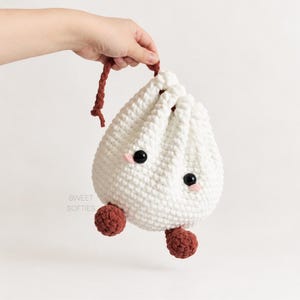 Puede incluir: Una bolsa de ganchillo blanca con cordón en forma de bola de masa con dos pies redondos de color marrón oscuro, un cordón marrón oscuro, ojos de cuentas negras y pequeñas mejillas rosas bordadas. El texto SWEET SOFTIES está impreso en gris claro.