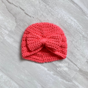 May include: A coral pink crocheted turban hat with a large bow on top. The hat is made with a soft yarn and has a comfortable fit.