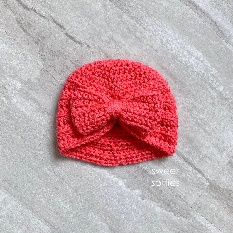 May include: A coral pink crocheted turban hat with a large bow on top. The hat is made with a soft yarn and has a comfortable fit.