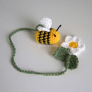 Könnte beinhalten: Eine gehäkelte Biene mit gelben und schwarzen Streifen, weißen Flügeln und einer schwarzen Antenne. Eine weiße Gänseblümchen mit gelber Mitte und grünen Blättern befindet sich neben der Biene. Eine grüne gehäkelte Ranke erstreckt sich von der Biene. Der Text "@SWEETSOFTIES" ist sichtbar.