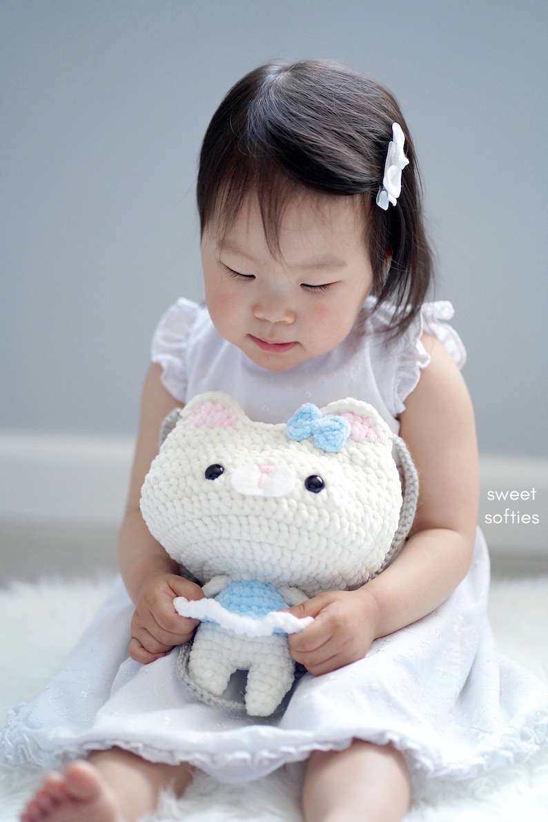 May include: A white crochet cat-shaped purse with a light blue bow and pink accents. The purse has black button eyes and a gray strap. A small, matching crochet doll is held inside. The image is labeled "sweet softies".