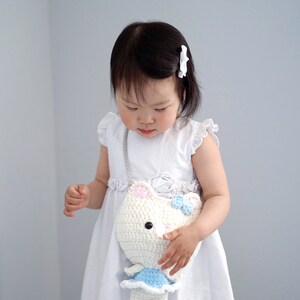 May include: A white crochet cat-shaped purse with pink ears and a blue bow. The purse has a light blue dress and a silver chain strap. The child is wearing a white dress and a white hair clip.