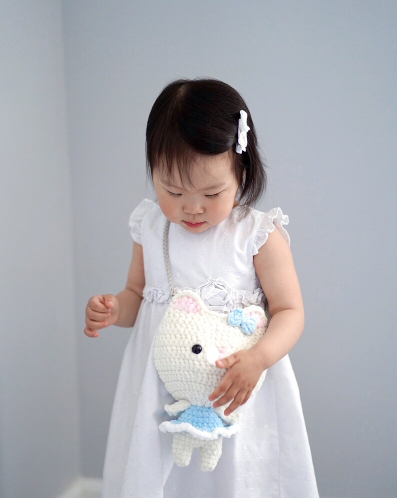 May include: A white crochet cat-shaped purse with pink ears and a blue bow. The purse has a light blue dress and a silver chain strap. The child is wearing a white dress and a white hair clip.