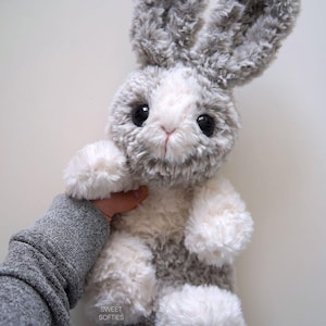 May include: A plush bunny rabbit with gray and white fur. The stuffed animal has large black eyes, a pink nose, and long, floppy ears. The bunny is soft and fluffy, with white paws and a gray body. The text "SWEET SOFTIES" is visible.