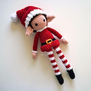 May include: A crocheted elf doll wearing a red and white Santa hat, red and white striped pants, and a red shirt with a yellow belt. The doll has a white pom-pom on the top of its hat and black shoes.
