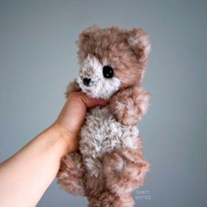 Op de afbeelding: Een kleine, bruine en witte teddybeer met zwarte ogen. De beer wordt vastgehouden door een hand.