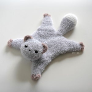 May include: A grey knitted plush toy shaped like a small animal with a long tail. The toy has a white tummy and brown ears. The toy is lying on its back with its legs and tail spread out. The text "SWEET SOFTIES" is printed on the toy's tail.