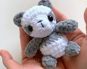 Può includere: Un piccolo orsacchiotto di peluche panda all'uncinetto con pelliccia grigia e bianca. Il panda ha occhi neri e un naso nero. Il giocattolo è tenuto nella mano di una persona.