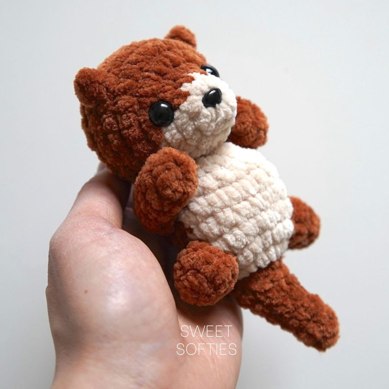 May include: A small brown and white crocheted stuffed animal resembling an otter. The animal has black eyes and a brown nose. The animal is being held in a person's hand.