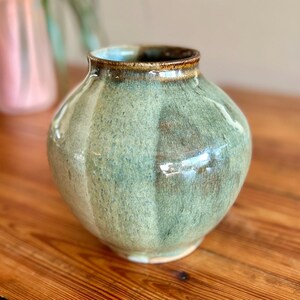 May include: A round ceramic vase with a wide body and a narrow opening. The surface features a mottled glaze in shades of sage green and light blue, with vertical indentations creating a faceted texture and a dark brown rim around the top edge.