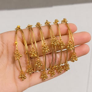 May include: A set of six thin gold-tone metal bangles, each featuring a delicate dangling charm composed of a cluster of small gold spheres. The bangles have a smooth, polished finish and are arranged in a parallel row to showcase the charms.