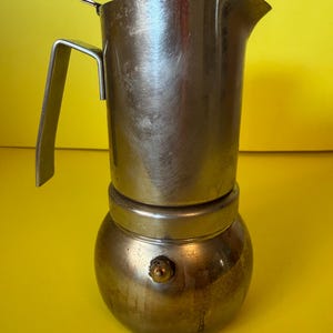 Puede incluir: Una cafetera de metal para estufa que presenta una cámara superior cilíndrica con pico vertedor, un mango plano y un depósito de agua inferior redondeado. El dispositivo tiene una válvula de liberación de presión en la base.