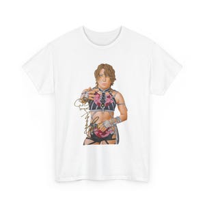 May include: A white short-sleeve t-shirt featuring a graphic print of a person with light brown hair wearing a black and white striped harness top with pink rose accents. The graphic includes a gold-colored signature overlaying the torso area.