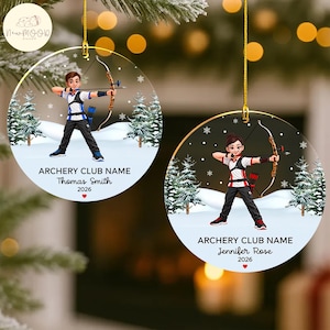 May include: Two round ceramic ornaments featuring cartoon archers in a snowy forest. One shows a person in a blue and white uniform, the other in red and white. Both include text reading ARCHERY CLUB NAME, names, and the year 2026.