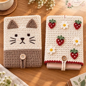 May include: Two rectangular crochet book covers. The left cover is cream and brown with a cat face design, featuring two ears, eyes, and whiskers. The right cover is cream with red strawberry and white daisy appliques. Both have a wooden button closure.