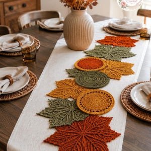 May include: A table runner featuring a series of crocheted autumn leaf motifs and circular coasters in shades of burnt orange, mustard yellow, and olive green. The runner is arranged on a wooden table with a logo reading BloomRunner Crochet.