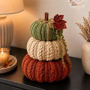 May include: A stack of three crocheted pumpkins in green, cream, and burnt orange. The top green pumpkin features a cinnamon stick stem, a twine bow, and a crocheted red leaf. The cream and orange pumpkins have a textured cable knit pattern.