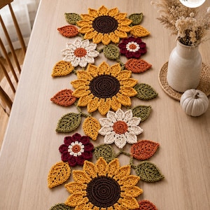 Puede incluir: Un camino de mesa de ganchillo con un patrón repetitivo de girasoles amarillos con centros marrón oscuro, margaritas color crema con centros naranjas, flores rojo oscuro y hojas verdes y naranjas unidas por tallos verdes.