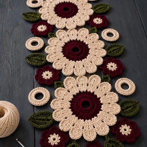 May include: A crochet table runner featuring large cream-coloured flowers with dark burgundy centres, smaller burgundy flowers with cream centres, and green leaves. The runner includes cream-coloured rings and a 3.5 mm crochet hook.