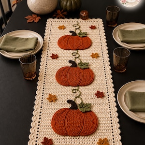 Peut inclure: Un chemin de table rectangulaire en crochet couleur crème orné de trois grandes citrouilles orange avec des tiges marron foncé et des feuilles vertes, agrémenté de petites feuilles d'automne crochetées en orange, jaune et rouge.