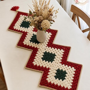 May include: A zigzag-shaped crochet table runner featuring a repeating pattern of dark green and cream-colored granny squares bordered by deep red yarn. The edges are trimmed with gold thread, and each end is finished with a red tassel.