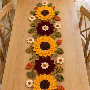 May include: A long crochet table runner featuring a repeating pattern of large yellow sunflowers with dark brown centers, deep burgundy dahlias, small white flowers, and green and orange leaves. The text BloomRunner Crochet is in the corner.