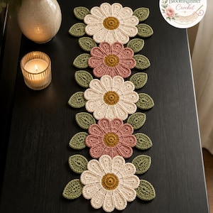 May include: A crochet table runner featuring alternating cream and dusty pink daisy-shaped flowers with mustard yellow centers, accented by small green leaves. The design includes a circular logo in the corner reading BloomRunner Crochet.