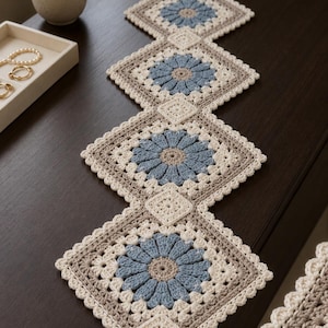 May include: A long, rectangular crochet table runner featuring a repeating pattern of square motifs. Each square contains a central blue flower with beige centers, surrounded by cream and beige yarn. The runner ends with a light blue tassel.