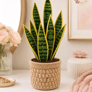 Op de afbeelding: Een gehaakte sansevieria met gevarieerde donkergroene en lichtgroene bladeren met felgele randen, geplaatst in een gestructureerde beige gehaakte mand. De plant heeft meerdere rechtopstaande, puntige bladeren en een bruine bodem.