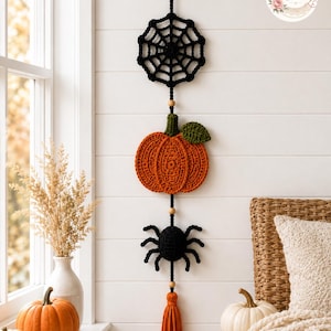 May include: A vertical crochet wall hanging featuring a black spider web at the top, a central orange pumpkin with a green leaf, and a black spider at the bottom. The elements are connected by black cord with wooden beads and an orange tassel.