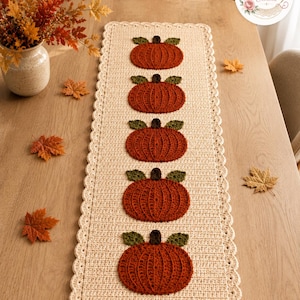 May include: A rectangular cream-colored crochet table runner featuring a scalloped edge and five evenly spaced, textured orange pumpkins with dark brown stems and dark green leaves appliqued in a vertical row along the center of the runner.