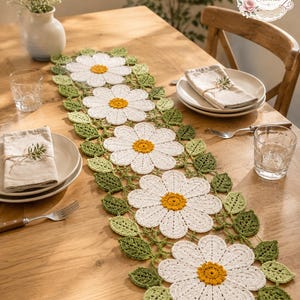 May include: A crochet table runner featuring a repeating pattern of five large white daisy flowers with yellow centers, surrounded by interconnected green leaves. The runner is laid out on a wooden table, with a logo reading BloomRunner Crochet.
