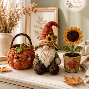 Puede incluir: Un conjunto de ganchillo que incluye una cesta de calabaza naranja con cara sonriente, un gnomo con sombrero color óxido con un girasol y un girasol en una maceta color canela con un corazón rojo. Los objetos están hechos de hilo y texturizados.