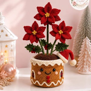 Könnte beinhalten: Ein gehäkelter Topf im Lebkuchen-Stil mit lächelndem Gesicht, der drei rote Weihnachtssterne mit gelben Mitten und grünen Blättern enthält. Der Topf hat einen weißen Zuckerguss-Rand, zwei rote Knöpfe und eine kleine rot-weiße Weihnachtsmütze.