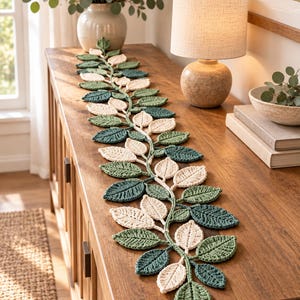 May include: A long, decorative table runner crafted from crocheted yarn, featuring a central vine design with alternating leaves in shades of cream, sage green, and dark forest green, arranged in a repeating pattern along the entire length.