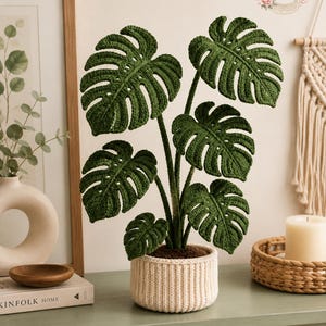 Könnte beinhalten: Eine gehäkelte Monstera-Pflanze mit tiefgrünen, geschlitzten Blättern und sichtbaren strukturierten Maschen. Die Pflanze steht in einem zylindrischen, gerippten, cremefarbenen Häkeltopf. Die Blätter sind an einzelnen Stielen angeordnet, die das natürliche Wachstum einer Monstera deliciosa nachahmen, wobei an der Oberseite des Topfes eine dunkelbraune, erdeähnliche Basis zu sehen ist.