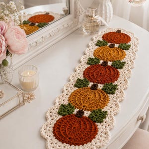 May include: A rectangular crochet table runner featuring five alternating orange and mustard yellow pumpkins with dark brown stems and green leaves, bordered by a cream-colored scalloped edge. The design includes the text BloomRunner Crochet.