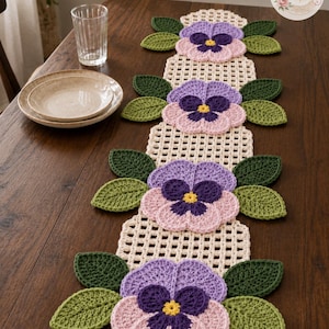 May include: A cream-colored crochet table runner featuring four pansy flower motifs in shades of light purple, dark purple, and yellow, accented by dark green crocheted leaves. The runner has a grid-like pattern and includes a BloomRunner Crochet logo.