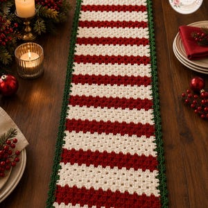 Puede incluir: Un camino de mesa rectangular de ganchillo con rayas horizontales alternas de hilo rojo oscuro y blanco roto. El camino está rematado con un borde de ganchillo de color verde oscuro sólido que enmarca toda la pieza, creando un borde texturizado.