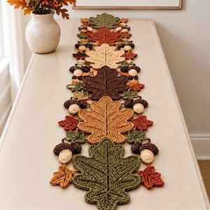 Op de afbeelding: Een gehaakte tafelloper met een herhalend patroon van herfstachtige eikenbladeren en eikels in tinten groen, bruin, oranje en crème. Het textielstuk is opgebouwd uit individuele gehaakte motieven die aan elkaar zijn gezet tot een lange strook.