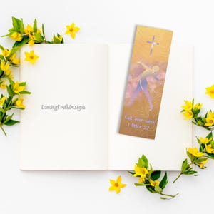 Könnte beinhalten: Ein rechteckiges Lesezeichen mit goldenem Farbverlauf, das ein Kreuzsymbol, die Silhouette einer nach oben reichenden Person und den Text "Cast your cares 1 Peter 5:7" in hellvioletter Schrift zeigt, platziert auf einem offenen Buch.
