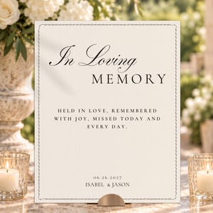 May include: A rectangular cream-colored memorial sign featuring a decorative border and black text that reads In Loving MEMORY, HELD IN LOVE, REMEMBERED WITH JOY, MISSED TODAY AND EVERY DAY, 06.26.2027, and ISABEL & JASON, supported by a small metal stand.