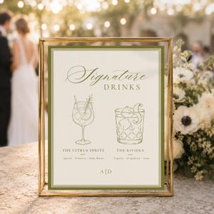 May include: A rectangular sign in a gold metal frame featuring the text Signature DRINKS. It displays line art of two cocktails, The Citrus Spritz and The Riviera, with their ingredients listed below each, and the initials A|D at the bottom.