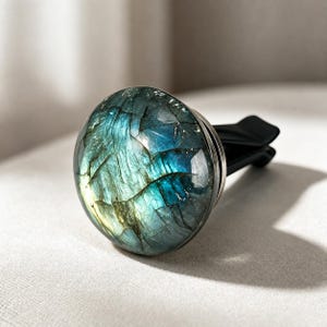 May include: A circular car air freshener vent clip featuring a polished labradorite stone face. The stone displays iridescent blue, green, and gold tones with natural internal fractures and a smooth, rounded surface set in a metal base.